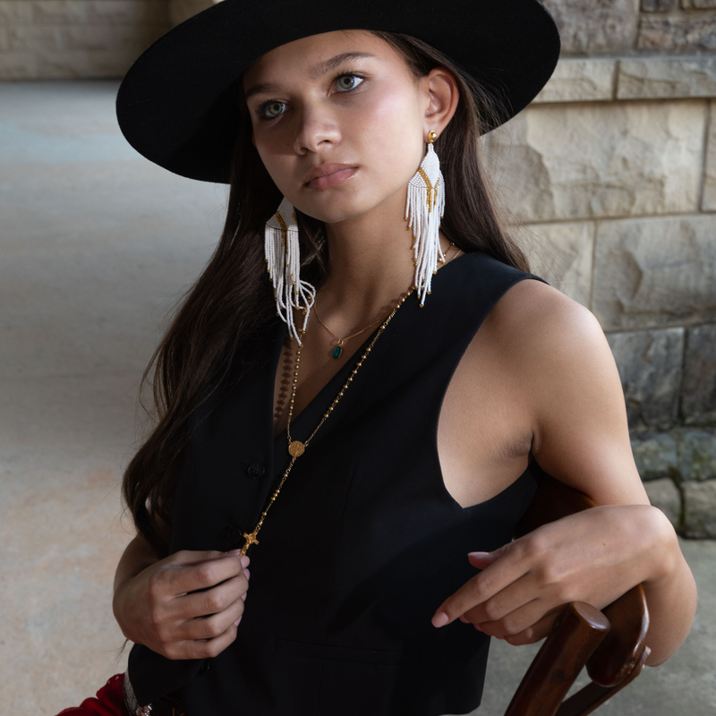 Woman wearing a black hat and black dress with Divine Goddess White Beaded earrings, standing against a stone wall.