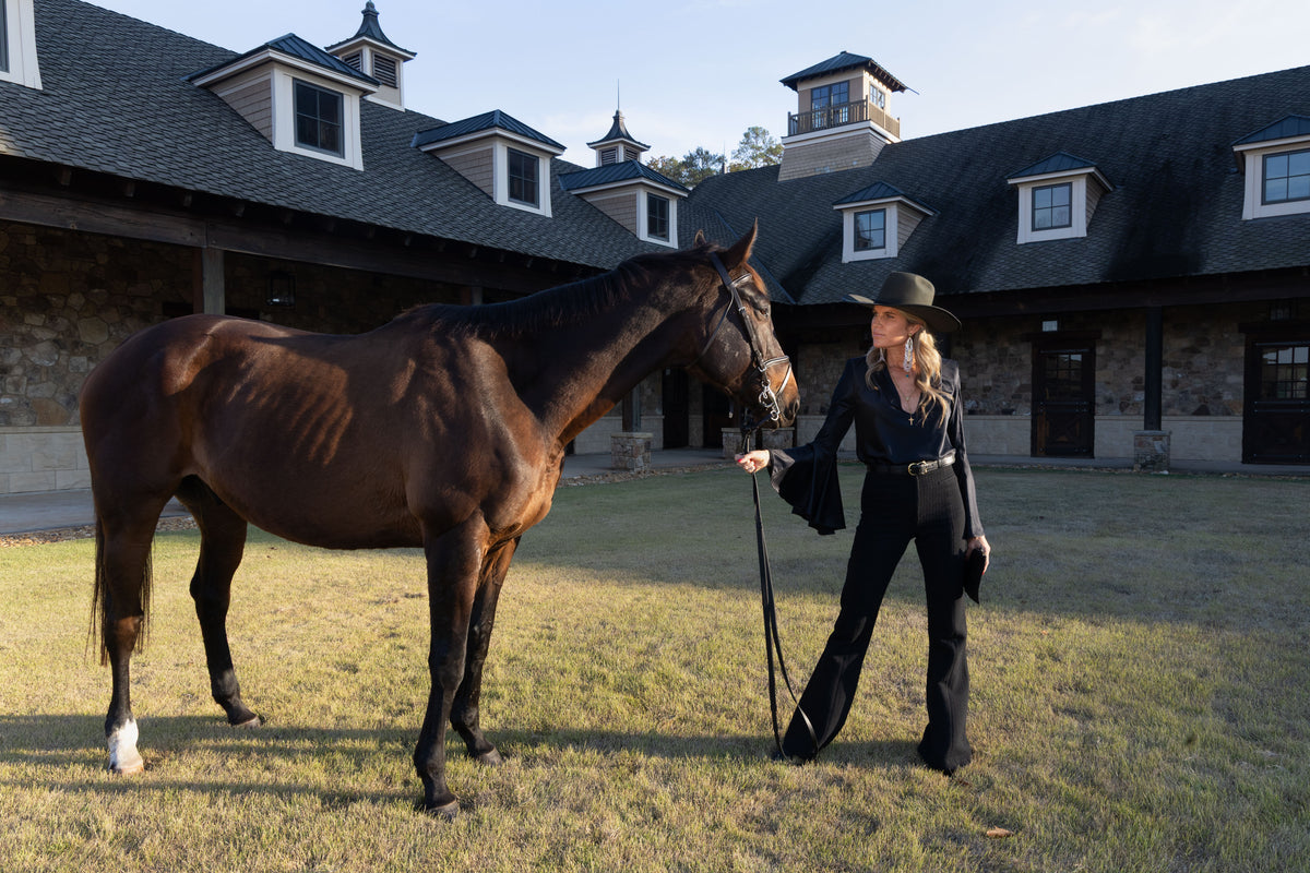 Woman in a cowboy hat walking a horse in front of a stable.